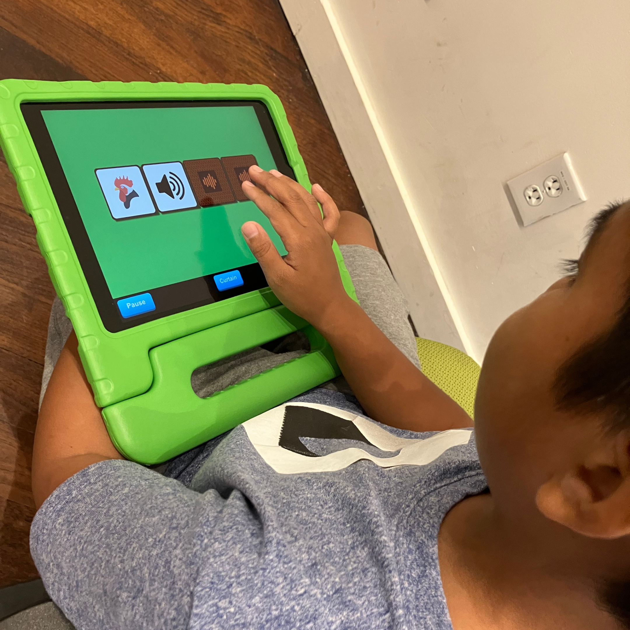 A young boy plays on a tablet. He is sitting on the ground, and the tablet is in his lap is in a protective green rubber case. The picture is an overhead view of the child tapping on the tablet screen and interacting with the FirstTap app