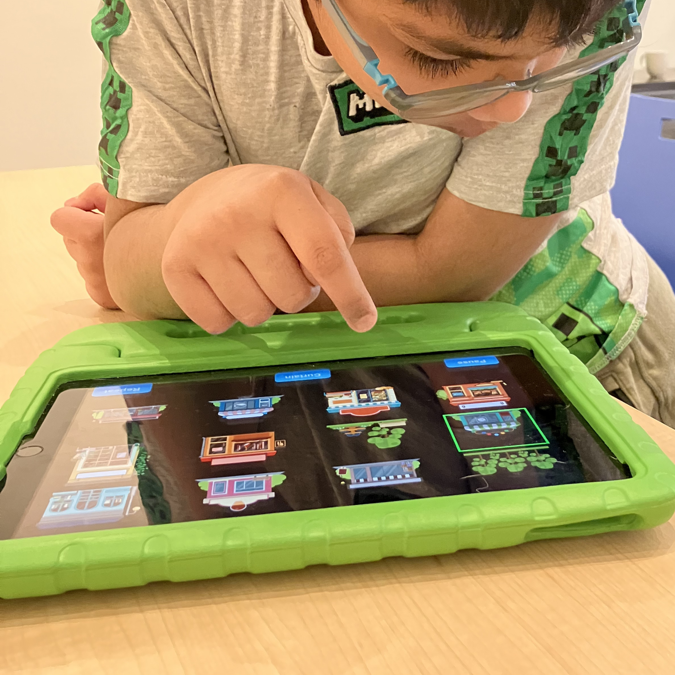 A young boy plays on a tablet. The tablet is sitting on top of a table in a protective green rubber case. The child tapping on the tablet screen and interacting with the FirstTap app.