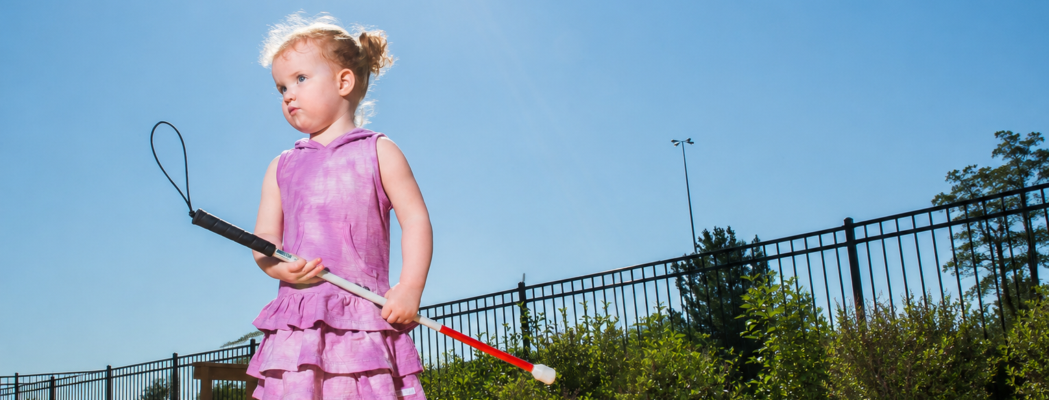 A young girl with light skin wearing a purple dress stands outside holding a small white cane