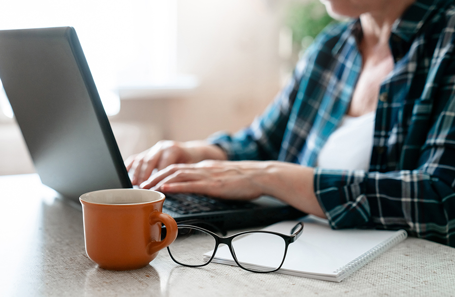 A person with light skin wearing a flannel shirt types on their laptop on a table - a mug, paper, and glasses are nearby
