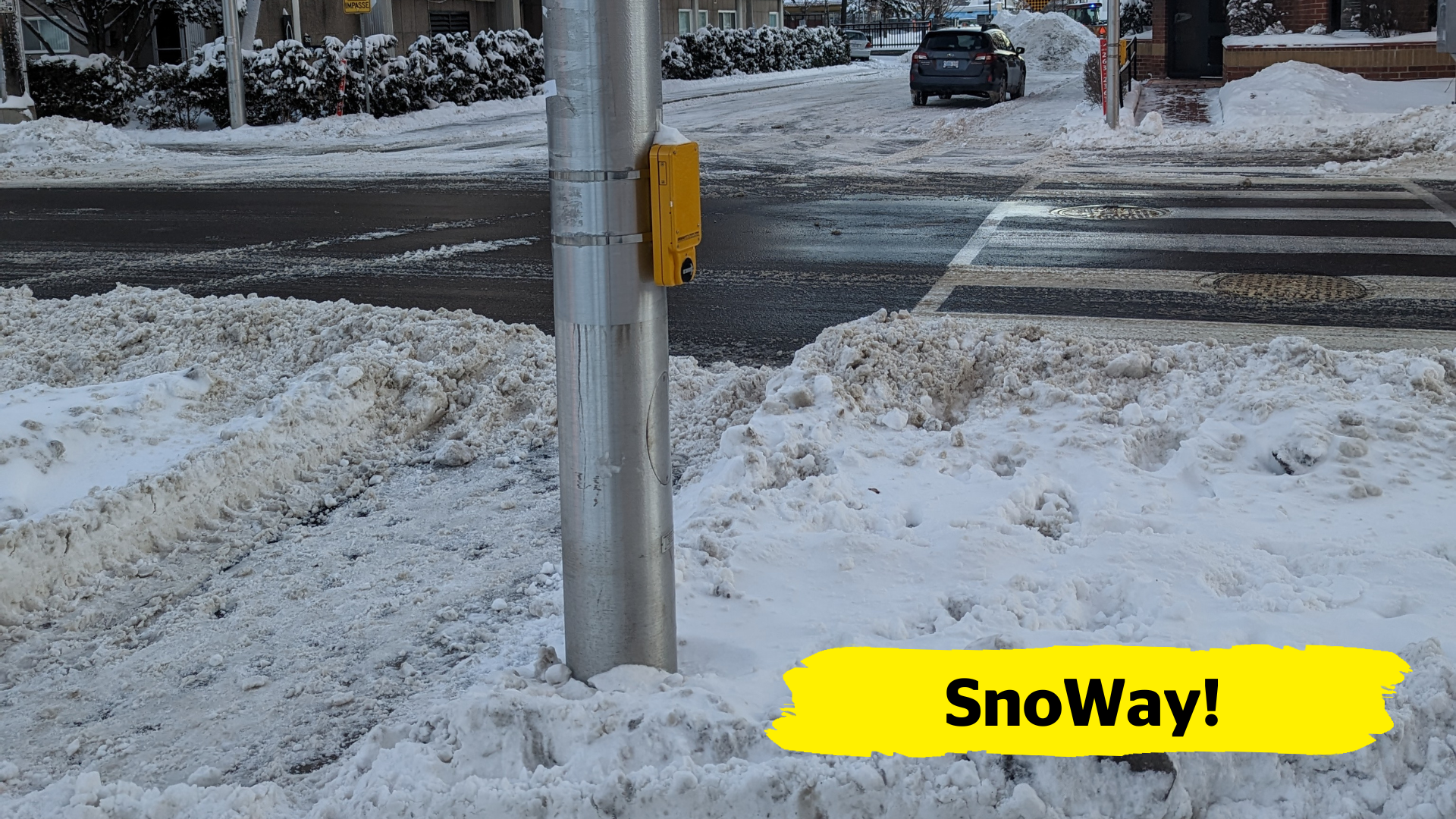 A pedestrian crosswalk is covered in snow and obstructing both the path of travel and a pole that houses the pedestrian crossing push-button.  In the bottom right-hand corner of the image, there is a yellow banner overlay with the text “SnoWay!”