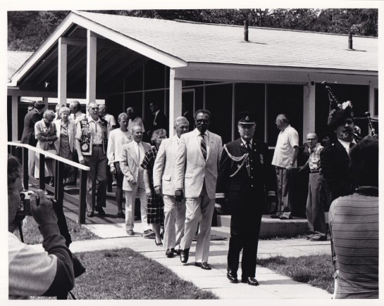 Photo en noir et blanc de Lincoln Alexander, lieutenant-gouverneur de l’Ontario, marchant derrière un joueur de cornemuse et un homme en uniforme au Centre Lake Joseph d’INCA le 20 juillet 1986.  