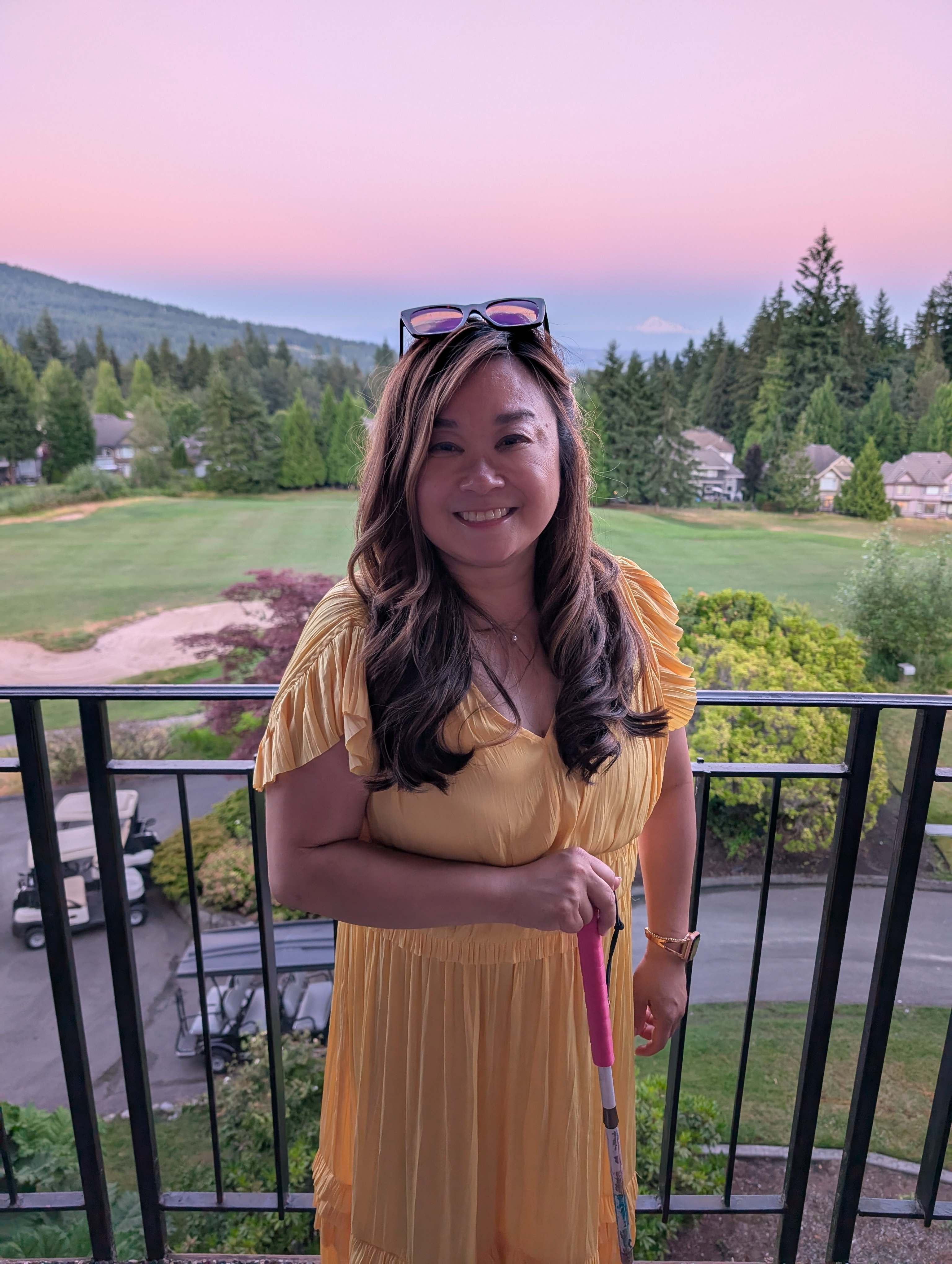 Anne Mok stands on a balcony in front of a sunset sky. She's wearing a yellow dress, has sunglasses on her head, and is holding a white cane.