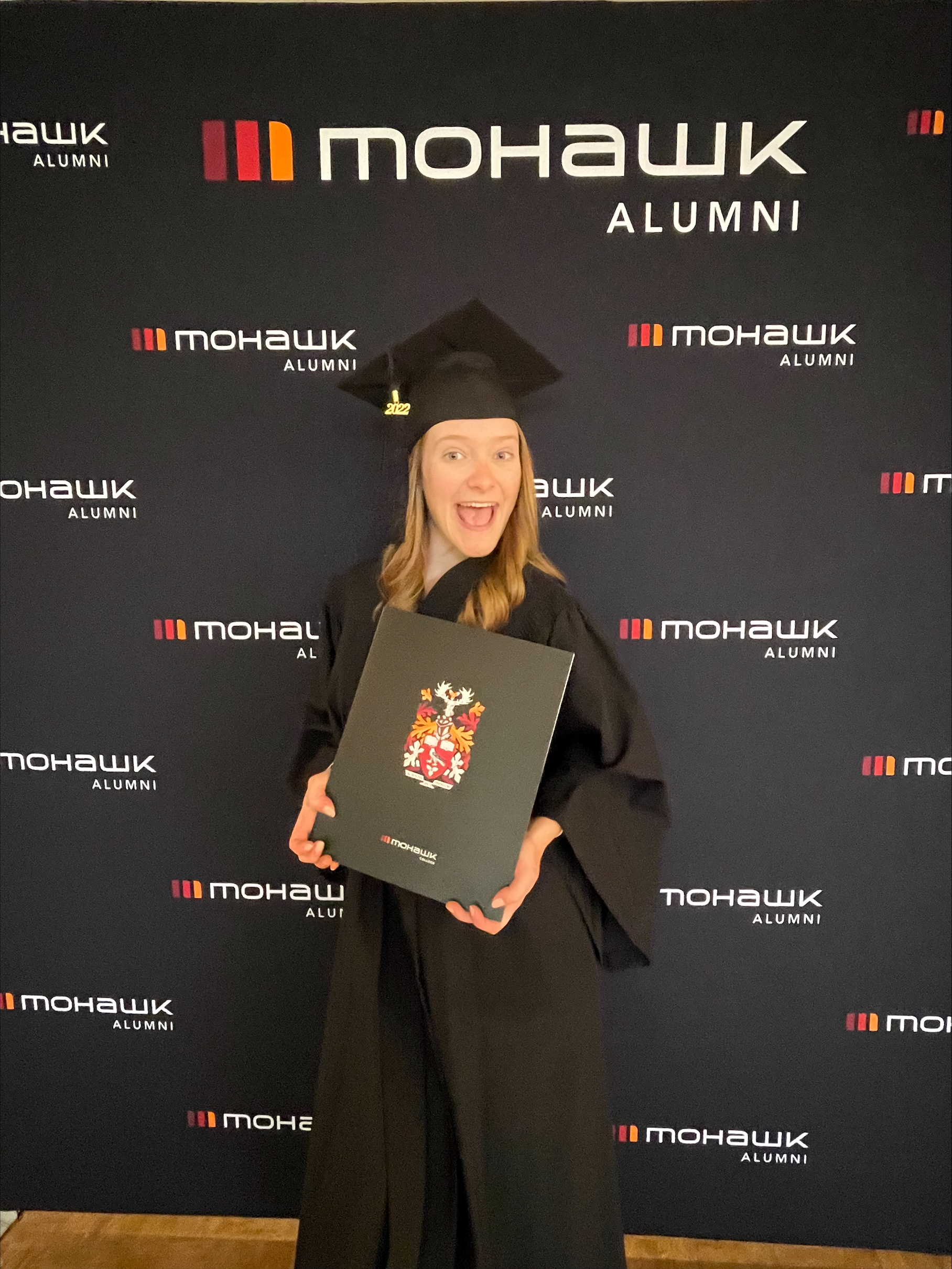 Graduation Day! Emilee, in her cap and gown, smiles and proudly displays her diploma. She is standing in front of a Mohawk College Alumni branded backdrop.