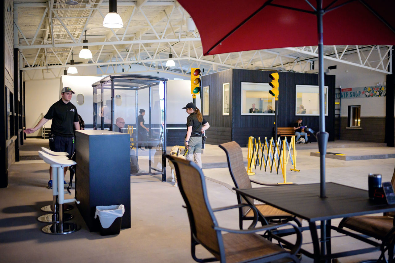 Wide shot of CNIB Guide Dogs’ Indoor City, a realistic training space styled like a cityscape. In the foreground, a patio-style coffee table with an umbrella is positioned near a high table with bar stools. Farther back are realistic training elements, including a bus shelter, sidewalks with curbs, safety barriers, traffic lights, and park benches. Several trainers walk future guide dogs through the brightly lit space.