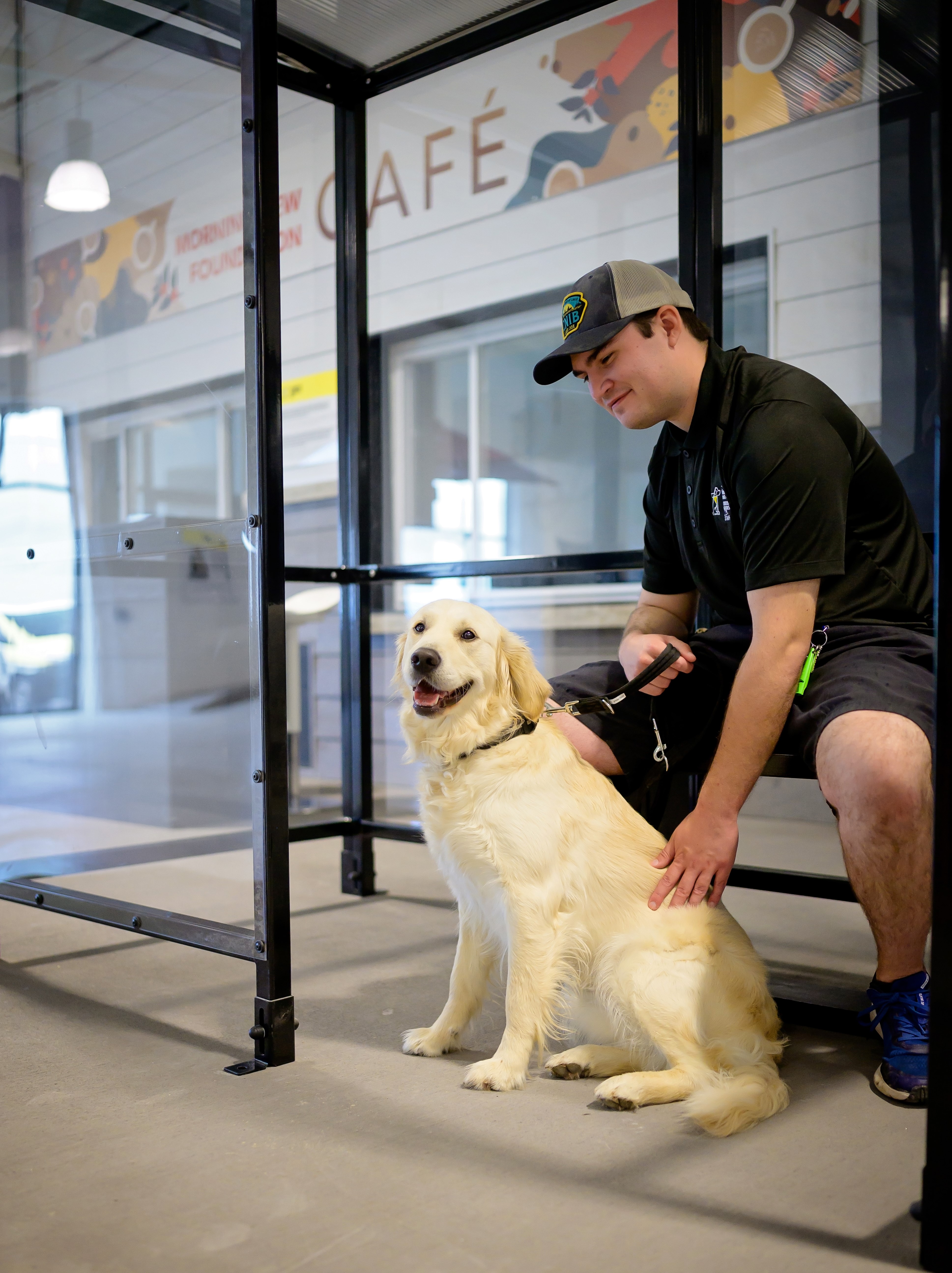 A smiling golden retriever sits beside her trainer, who wears a cap and black shirt, at a simulated bus stop inside CNIB Guide Dogs’ Indoor City. A storefront sign reading “Café” is visible in the background.