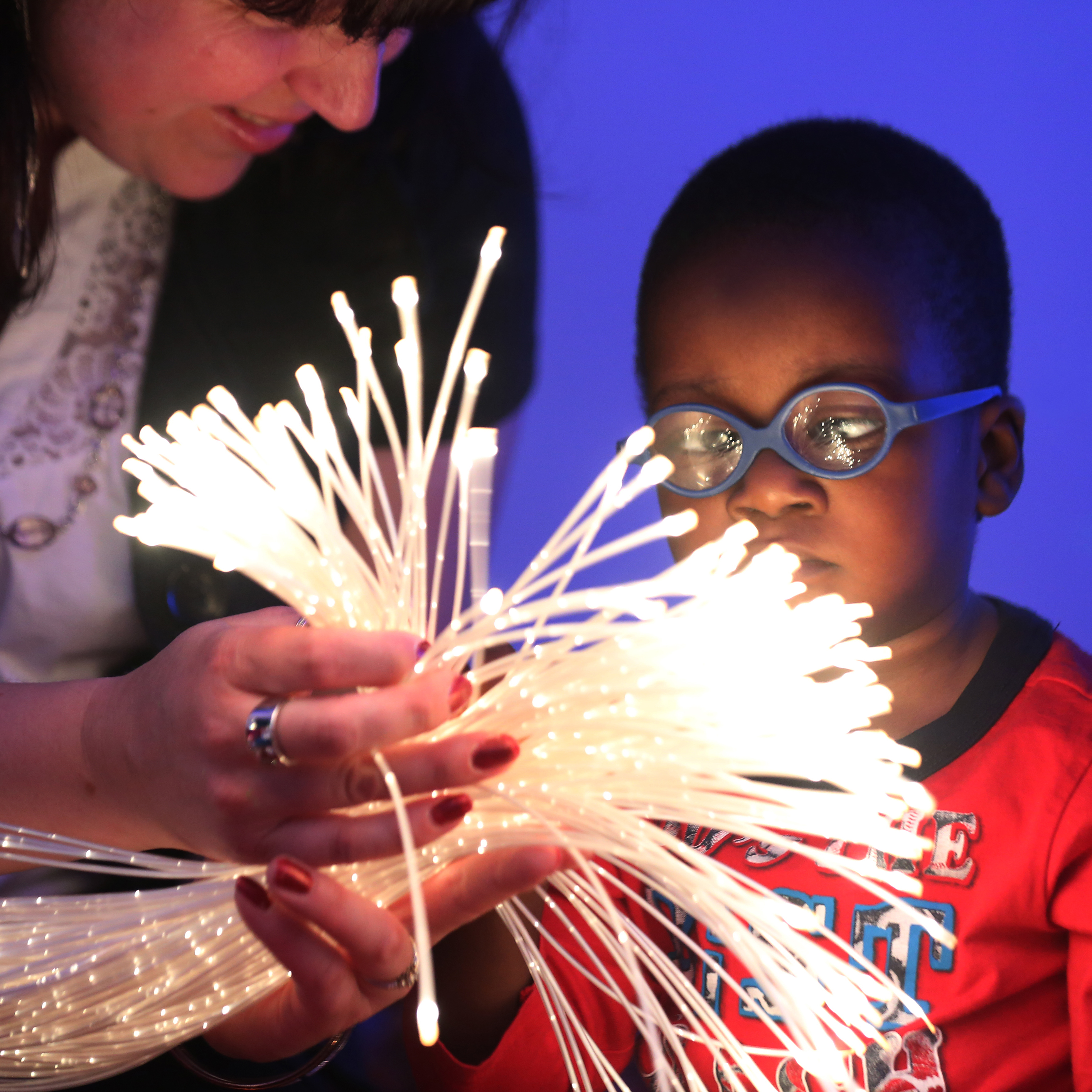 A toddler with dark skin and glasses is shown a light-up tactile toy by an adult