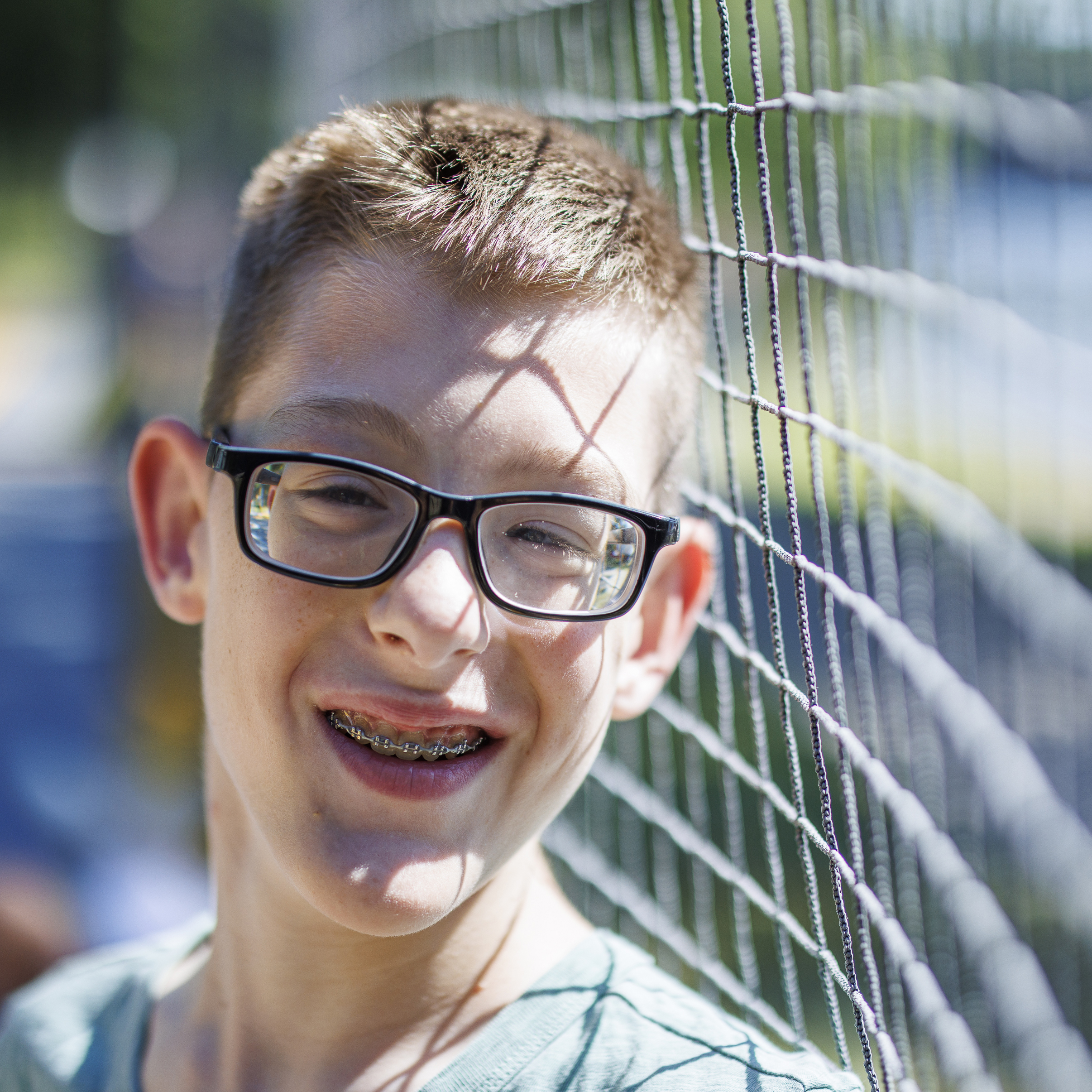 A boy with glasses and light skin smiles beside a volleyball net