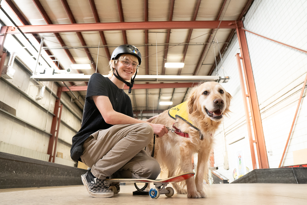 A young man with light skin, wearing a black helmet and glasses, smiles and kneels on a skateboard beside his golden retriever buddy dog. They are in an indoor skate park, and the dog wears a yellow CNIB vest.