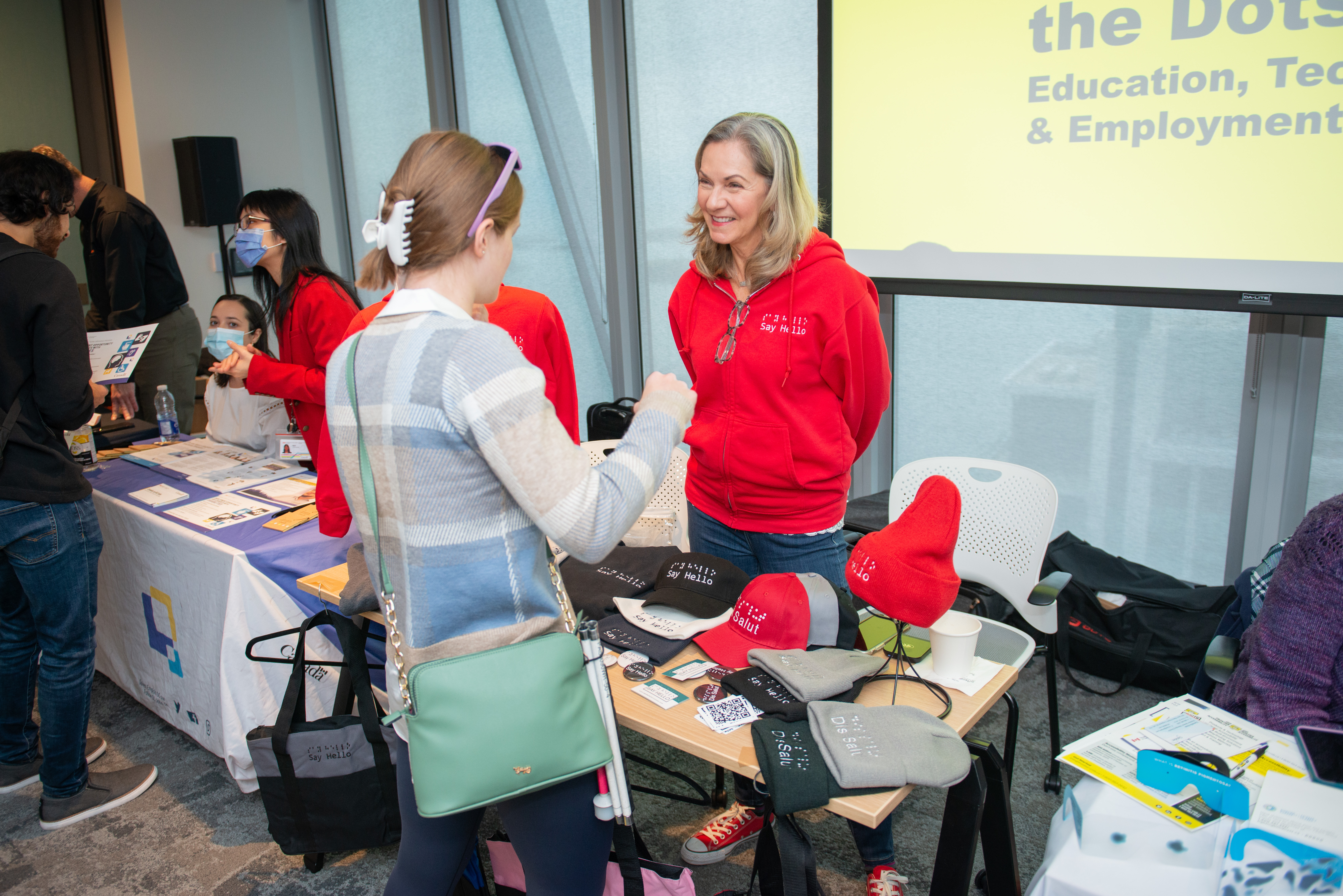 A woman wearing a red sweater stands at a booth selling shirts and hats at the connecting the dots conference in Toronto. Another woman looks at the table and talks with her.