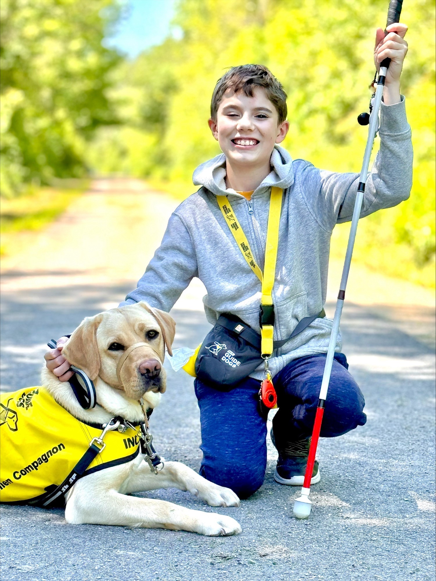 Caleb, a smiling boy with light skin, holds his white cane and crouches down next to Amber, a yellow Labrador retriever. Amber, lying down, looks at the camera with a serious expression while wearing her black and yellow CNIB Buddy Dog vest. They pose on a quiet paved road with green trees behind them.