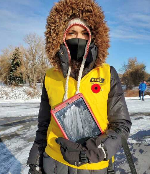 CNIB fundraiser in a parka and a bright yellow vest wearing a mask and holding a tablet