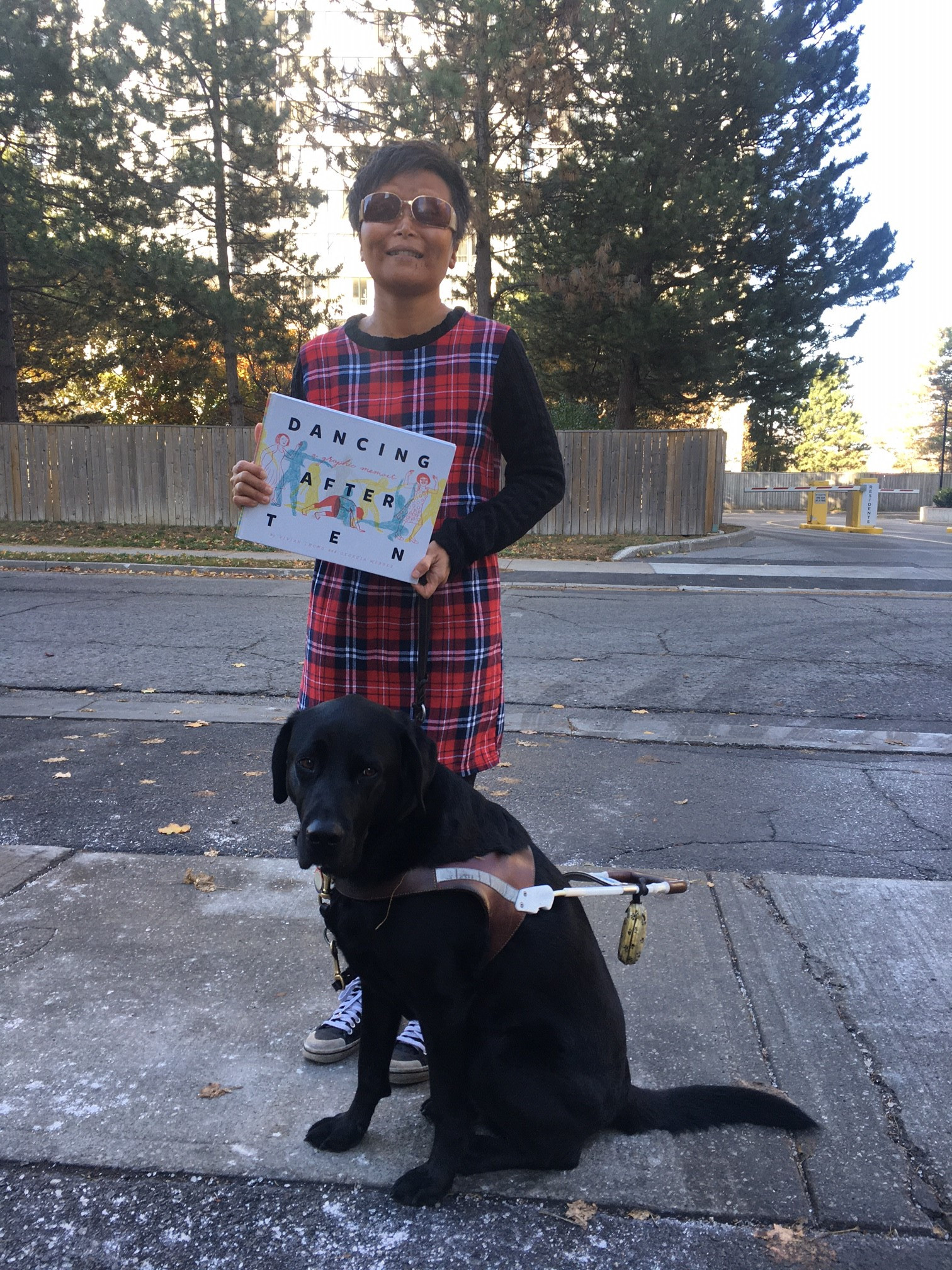 Vivian Chong stands outdoors and holds her book, Dancing after TEN. Her black guide dog, Catcher, stands at her feet. Vivian is wearing a plaid dress, sunglasses, and a big smile. 