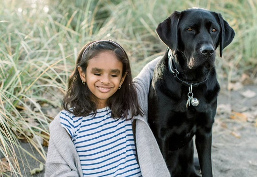 Deepa et Chelsey, un Labrador-Retriever noir, assis sur le sol devant une zone herbeuse; Deepa, toute souriante pour la photo, entoure son chien Chelsey de son bras