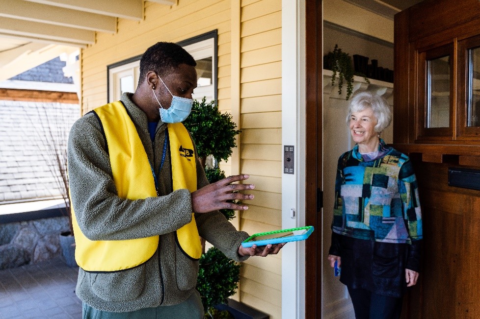 A CNIB Fundraiser in a yellow mask and vest, holding a tablet, speaking with a donor at her doorstep