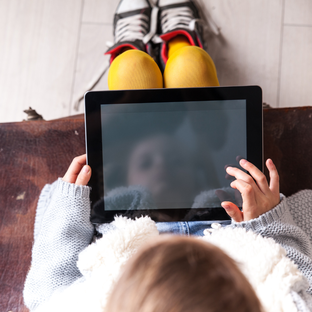 A young child sits on a couch and plays on a tablet. The picture is an overhead view of the child playing on the tablet and their reflection shows on the tablet screen.