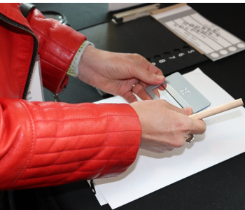  A woman holds a signature guide and a large grip pencil over a piece of paper. Beside her is a large print ballot and tactile voting template. 