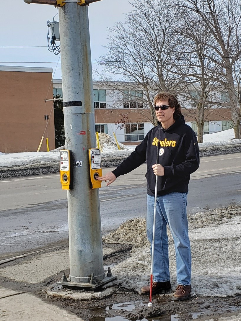 Bill Liggins se tient au coin de la rue à un panneau de signalisation piétonnière accessible. Il tient sa canne blanche dans la main gauche et appuie sur le bouton du passage pour piétons avec la main droite. 