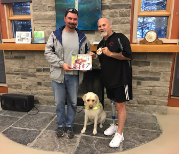 Guy Carriere & his guide dog (left) and Paul Rosen (right) pose for a photo together. Guy is holding an autographed picture of Paul and Paul is displaying a medal in his left hand. 