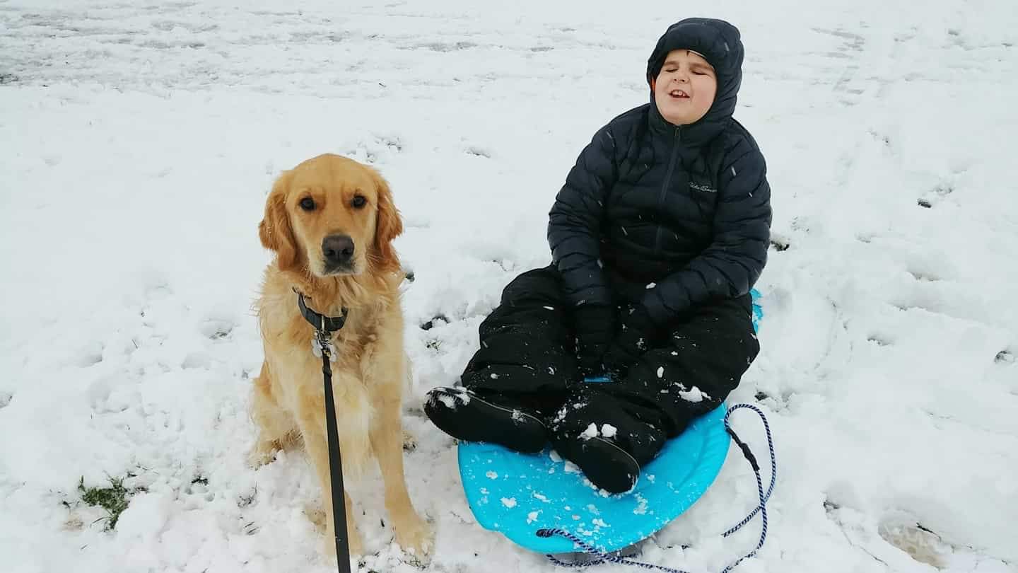 Landon and Ruggles, a golden retriever, sitting next to each other in the snow smiling for the camera; Landon is sitting on a sled and wearing a snowsuit.