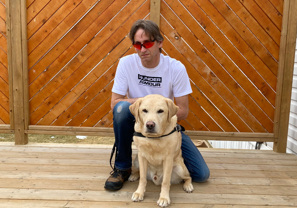 Gary Peasgood, standing outdoors against a wood patio wall with his guide dog. 