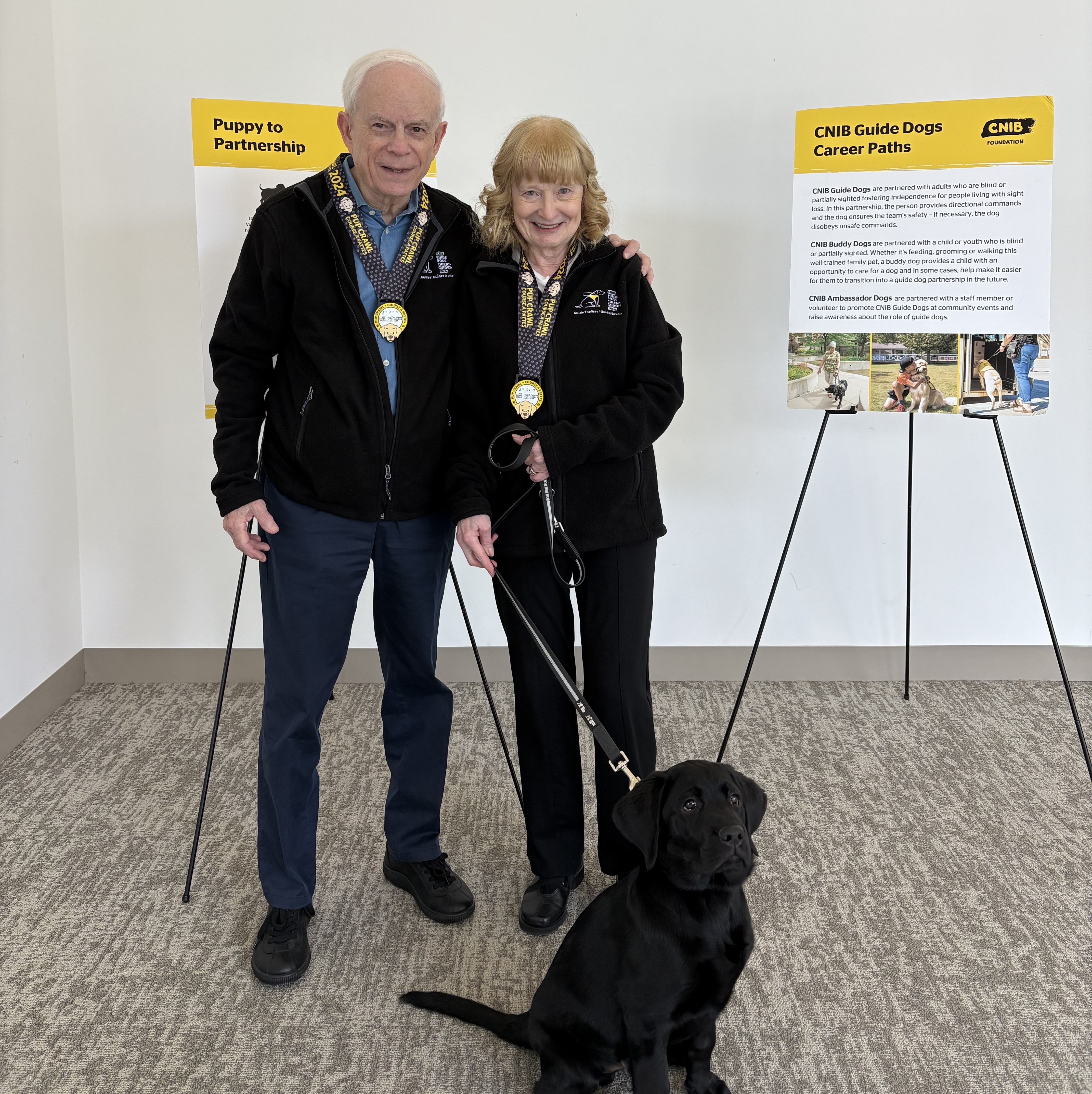 John and Mary Crocker, a couple with light skin, stand close together. John has his arm around Mary, who holds the leash of a black Labrador retriever puppy. Both wear Pup Crawl medals. Behind them are two posters – one says “Puppy to Partnership” and the other says “CNIB Guide Dogs Career Paths.”