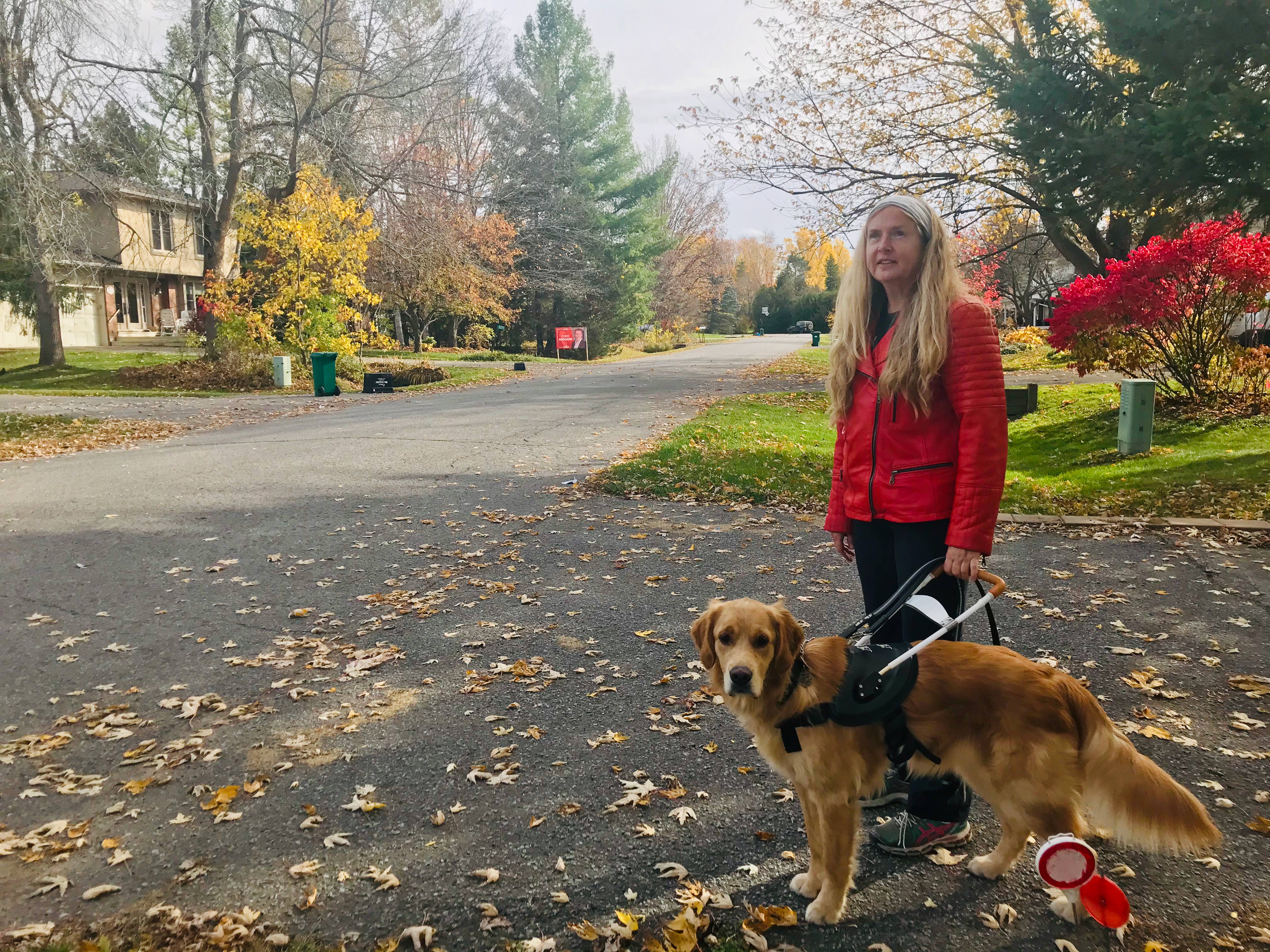 Diane, vêtue d’une veste rouge, est debout dans une rue couverte pleine de feuilles d'automne multicolores, tenant le harnais de Carla et souriant pour la pose. 
