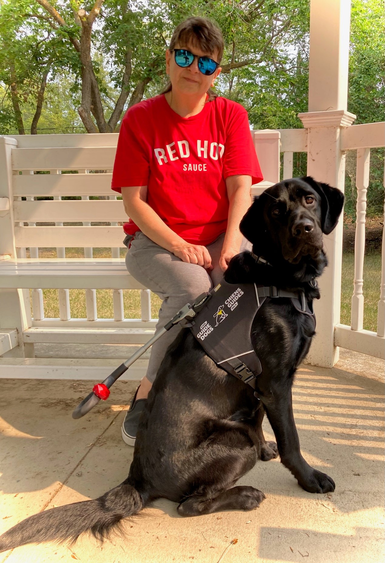 Jodi is sitting on a bench in a gazebo outdoors. Shadow, a black Labrador-golden-retriever mix, is sitting on the ground in front of her.