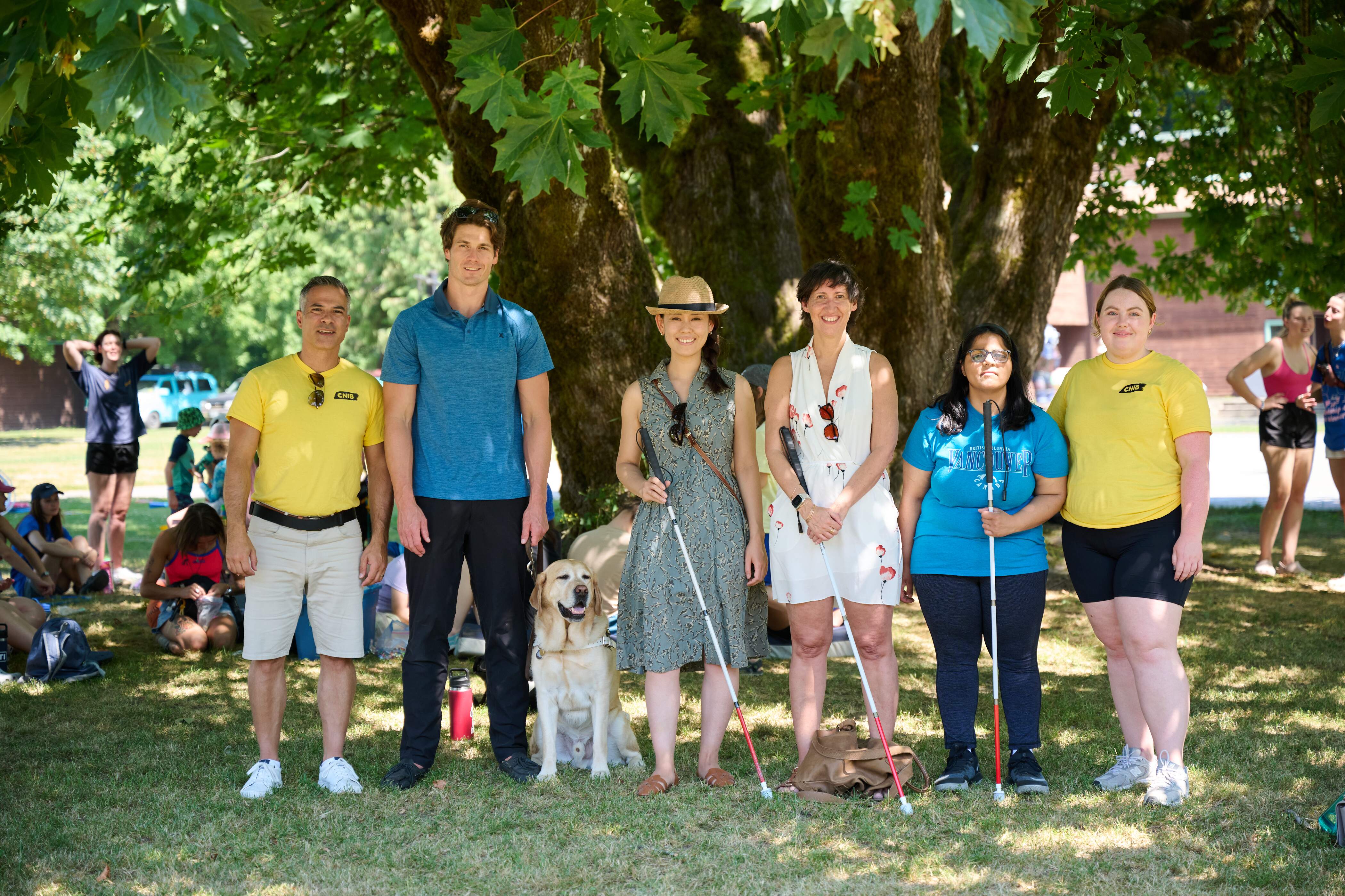 a group of people outside, some with white canes and guide dogs, others without. 