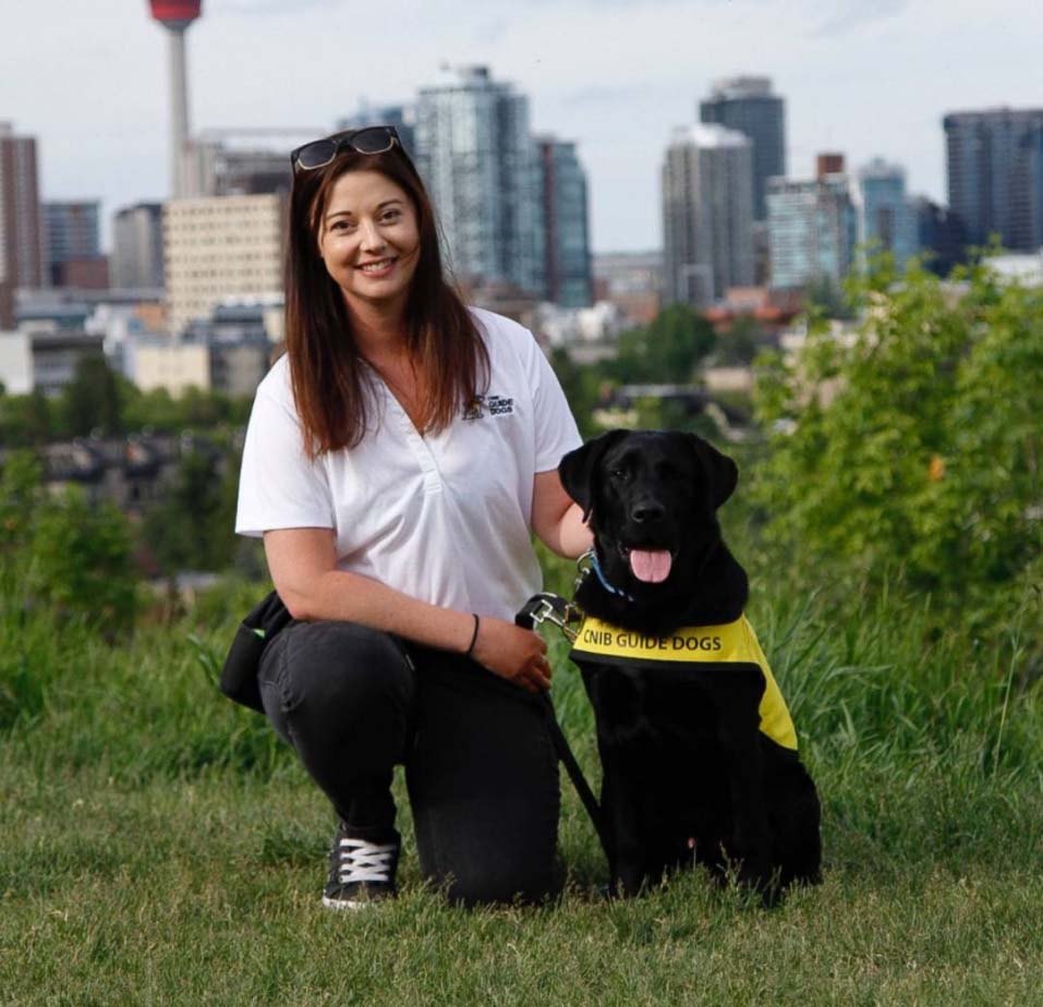 Kezia kneeling in the grass next to a future CNIB Guide Dog, smiling for the camera with a gorgeous cityscape in the background.