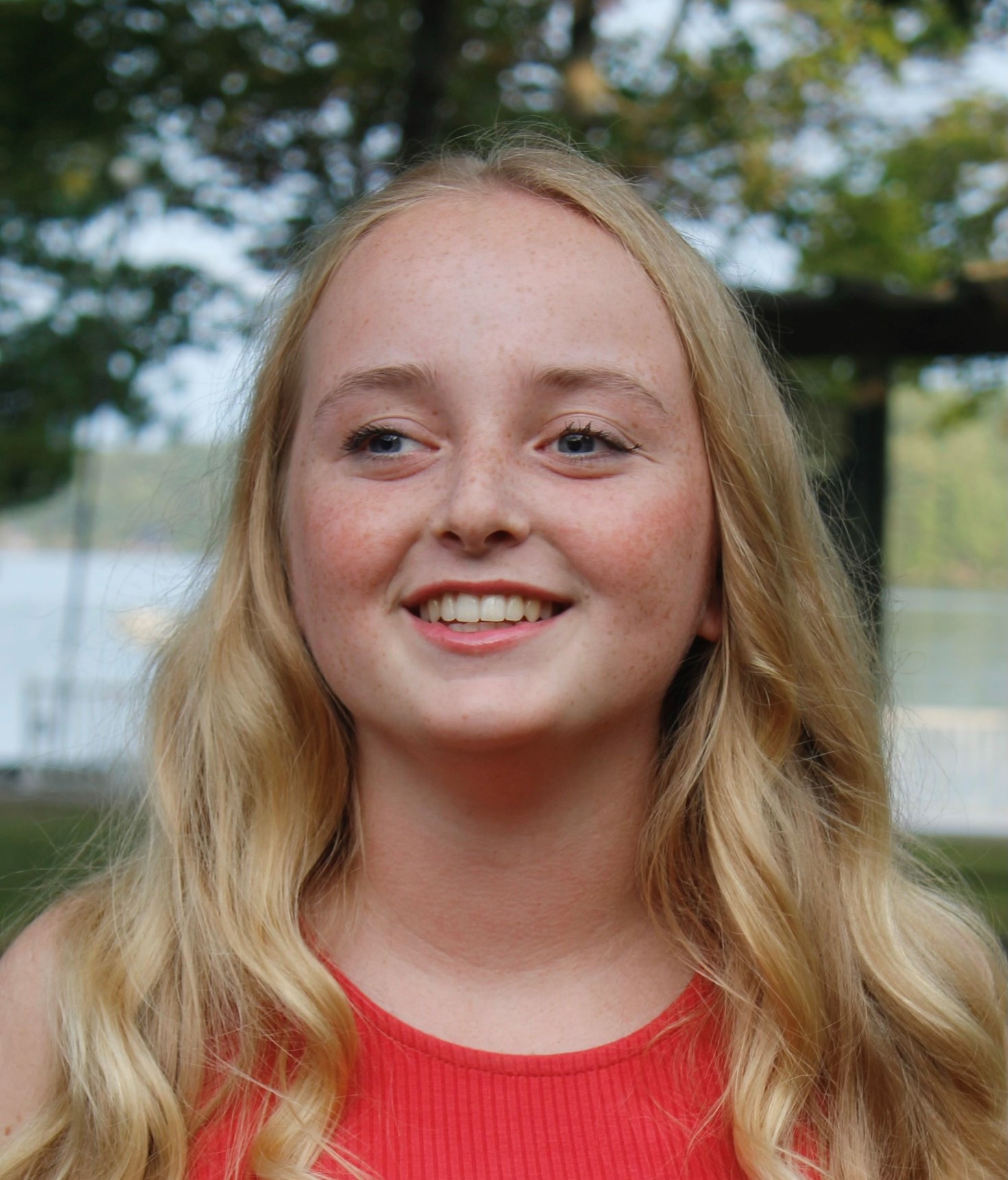 A young woman with long blonde hair is standing outside. She is wearing an orange shirt with trees and water behind her. She is smiling at the camera.