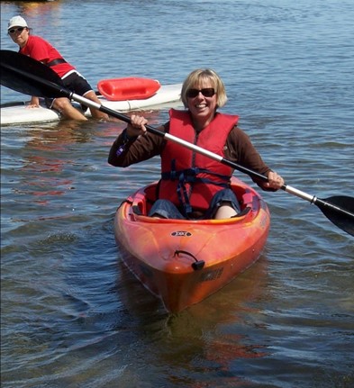 ​​Joanne smiles and poses in an orange kayak for a photo on the lake. She’s wearing sunglasses, a red life jacket, and holding her paddle. 