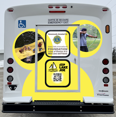 The back door of the new minibus. The door features a bright decal with abstract yellow bubble designs and photos of campers participating in archery and enjoying the lake. In the centre of the decal design is the text: “Generously donated by Lions Clubs International Foundation, Lions of District A12, MDA Lions and Friends” accompanied by the Lions Club International Fund logo, CNIB Lake Joe Logo, and a QR code.