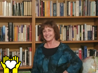 Lynn Kennedy poses for a photo in front of a book shelf. A yellow graphic of hands holding a white heart outlined in black in the bottom left corner