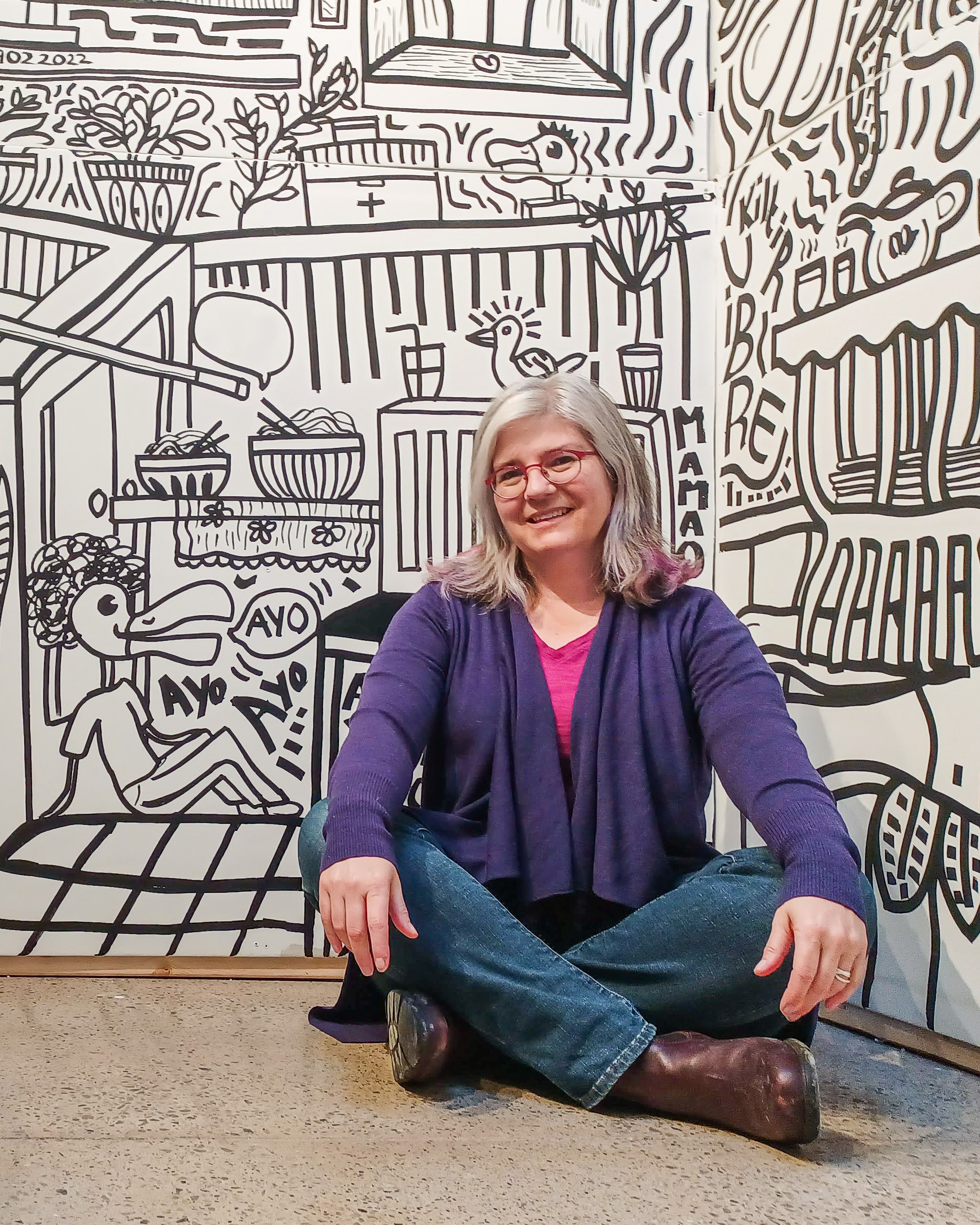 Mireille Messier smiles and sits on the floor in front of a painted mural. 
