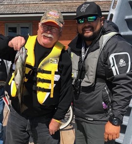 Bruce Roulston posant à côté de Eugene Chong avec un poisson pêché du quai sur la rive du Lake Joseph. Ils portent tous deux des gilets de sauvetage et une casquette. 