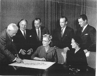 A group of men and women sitting and standing around a piece of paper pointing at it, circa 1960s. This photo is part of the opening program. This is the Lake Joseph Building-Planning Committee studying plans for the Adjustment-Training and Holiday Centre. The man on the left is Wilfred C. James (grandfather). The woman seated in front, centre is Miss Elsinore C. Burns (great aunt). 