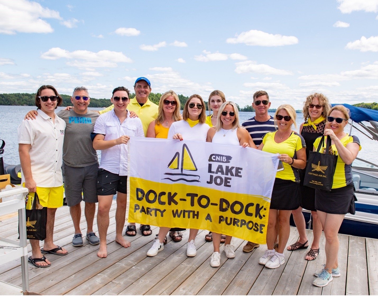  A group of event volunteers on the CNIB Lake Joe dock holding a "CNIB Lake Joe Dock-to-Dock: Party with a Purpose" flag