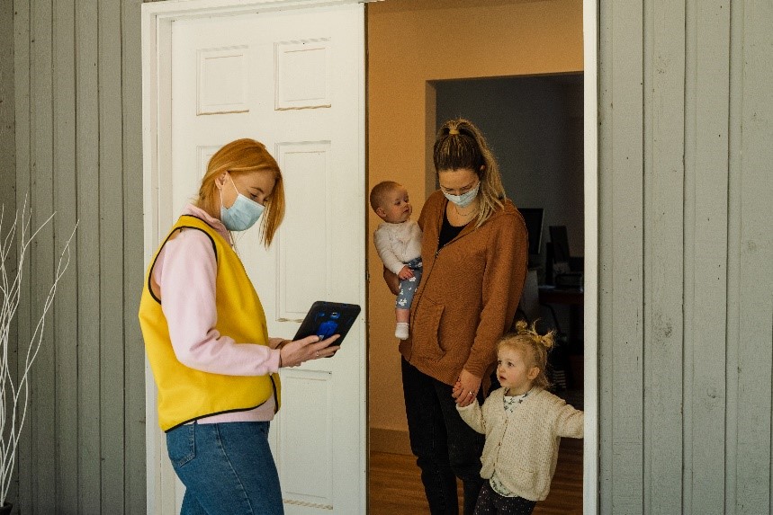 A CNIB Canvasser in a yellow vest and face mask, pointing to her tablet, while standing in an open doorway with a donor and the donor’s two small children.