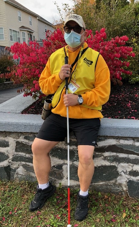 A CNIB Fundraiser in a yellow branded vest, ID badge and face mask standing with his white cane.