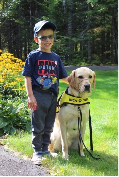Julien, un jeune garçon, est debout dehors à côté de Victor, son chien-compagnon, qui est assis à sa droite et porte un licou de contrôle « Gentle Leader ».