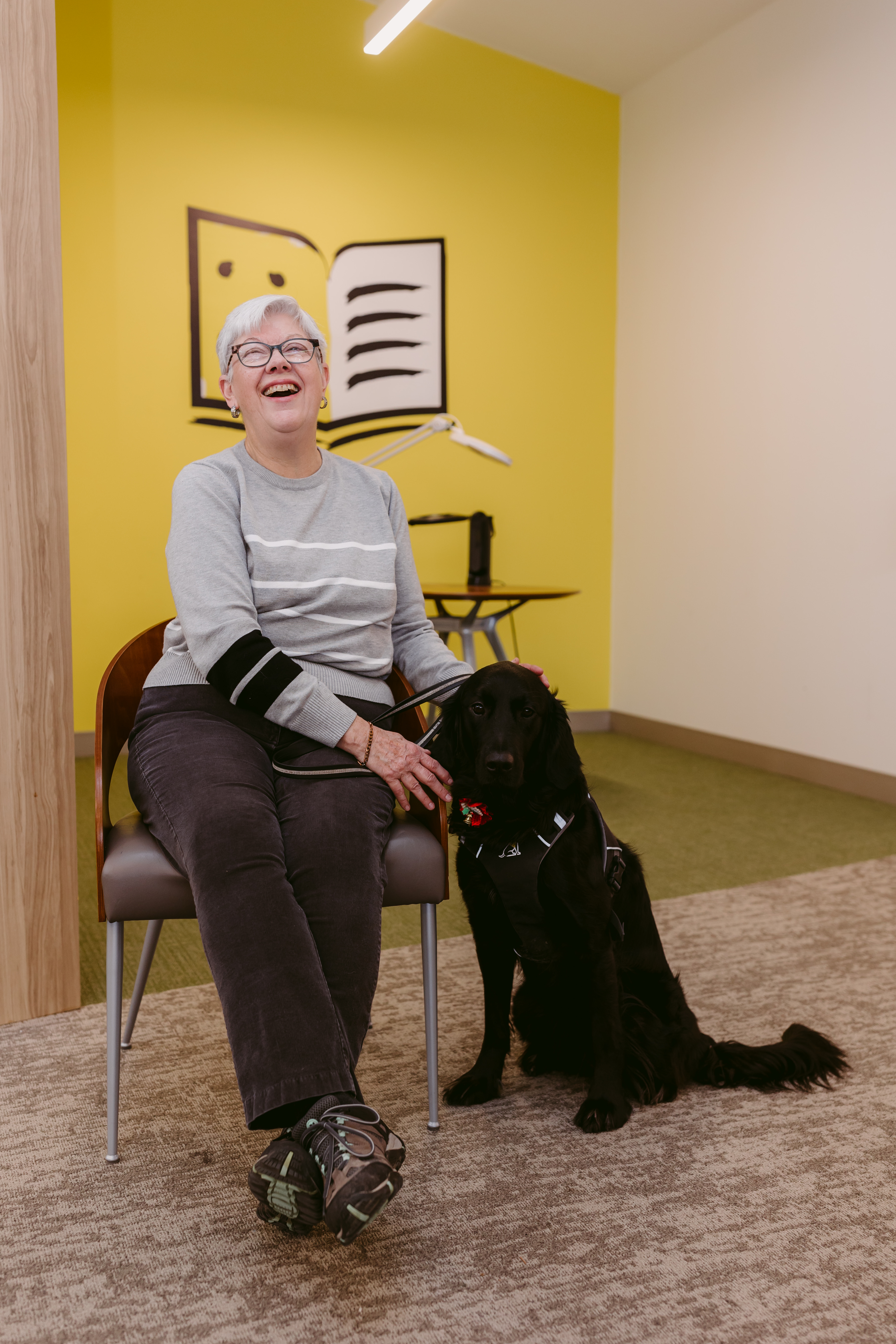 At a CNIB office, Penny sits in a chair. Her guide dog, Honour, a black Labrador retriever-golden retriever cross, sits at her feet. Penny pets Honours head and smiles with joy.