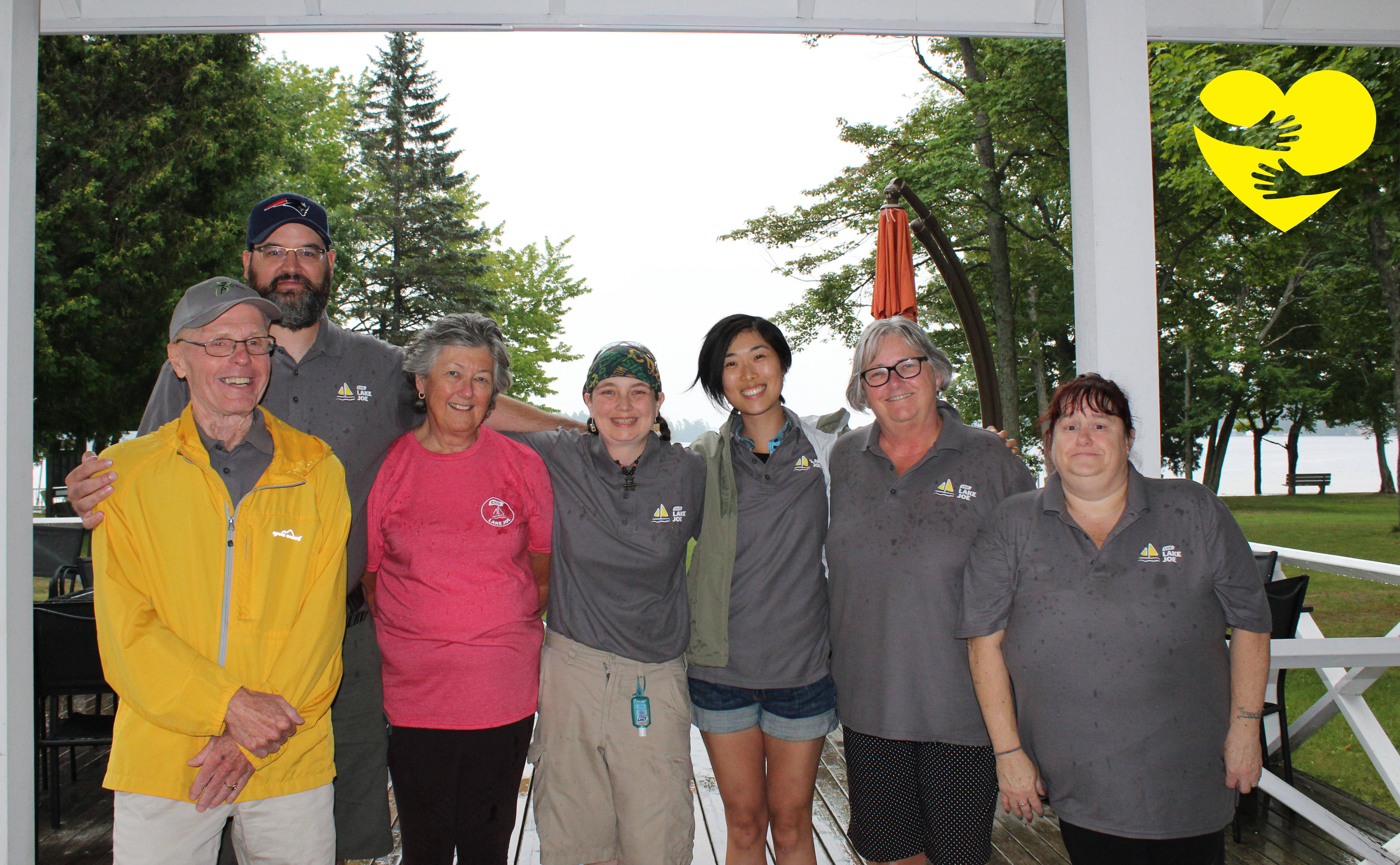 Sandra, wearing a pink shirt, smiling for the camera in a group photo with other CNIB Lake Joe volunteers. A graphic of arms hugging a cartoon yellow heart can be seen in the top-right corner of the photo.