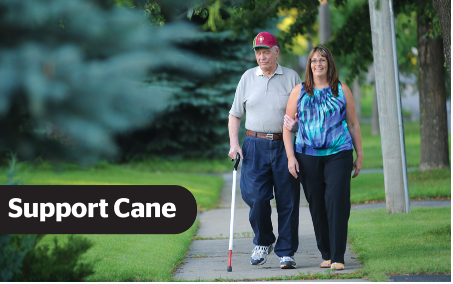 A man taking a walk with a woman along a paved path in a grassy area. In his right hand he is using a support cane and his left hand is holding onto the woman walking alongside him. The text "support cane" are overlayed in white text on a black background
