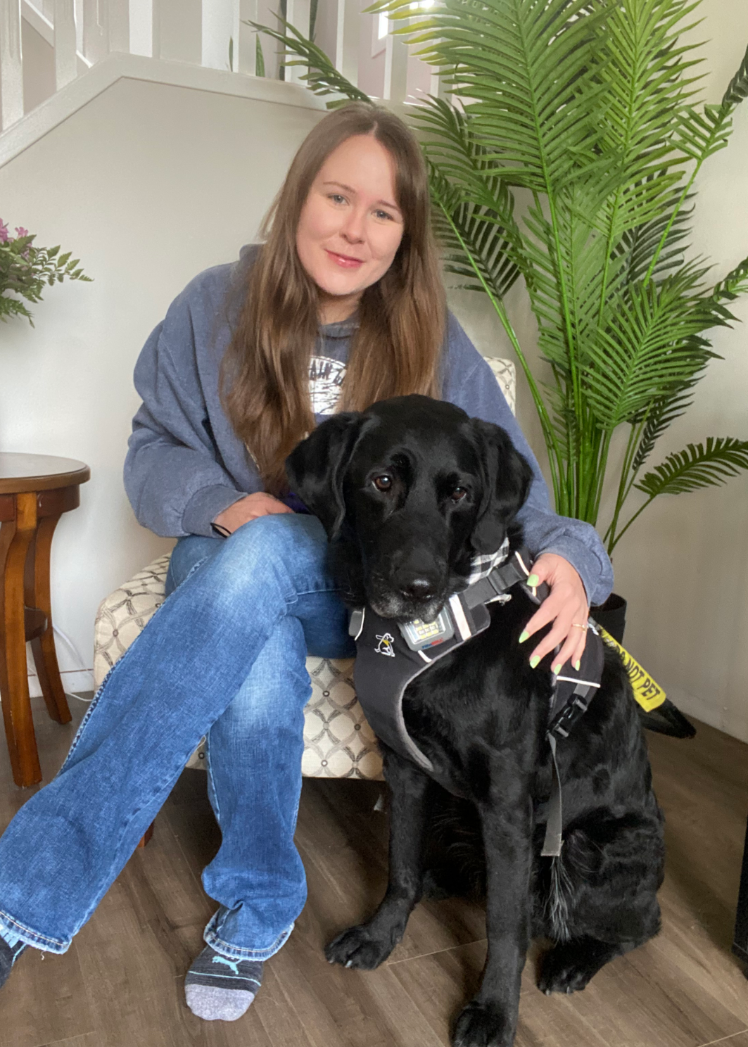 Taylor, a young woman with long brown hair, sits in a chair with her arm around her guide dog Wallace, a black lab who is wearing a harness and sitting beside her.