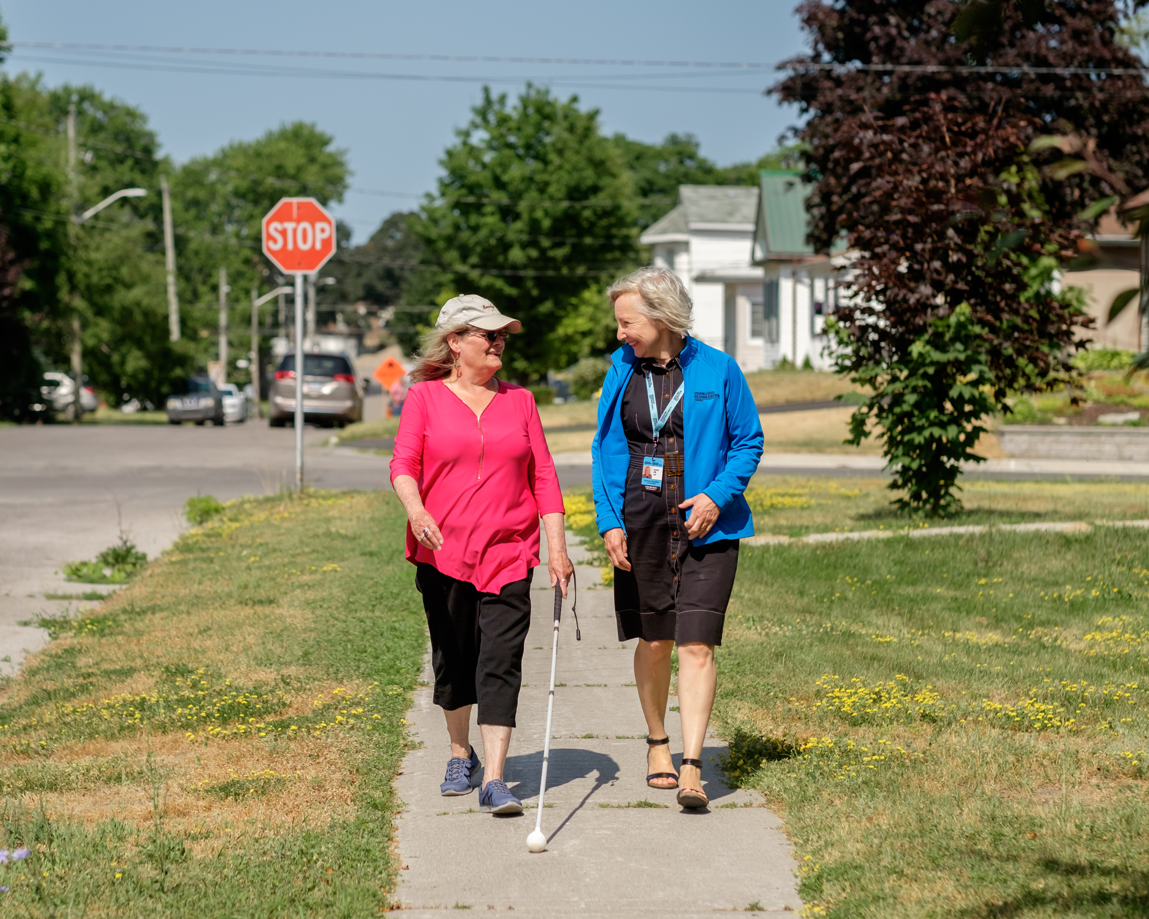 A VLRC Specialist walks beside a client using a white cane outside on a sunny day. 