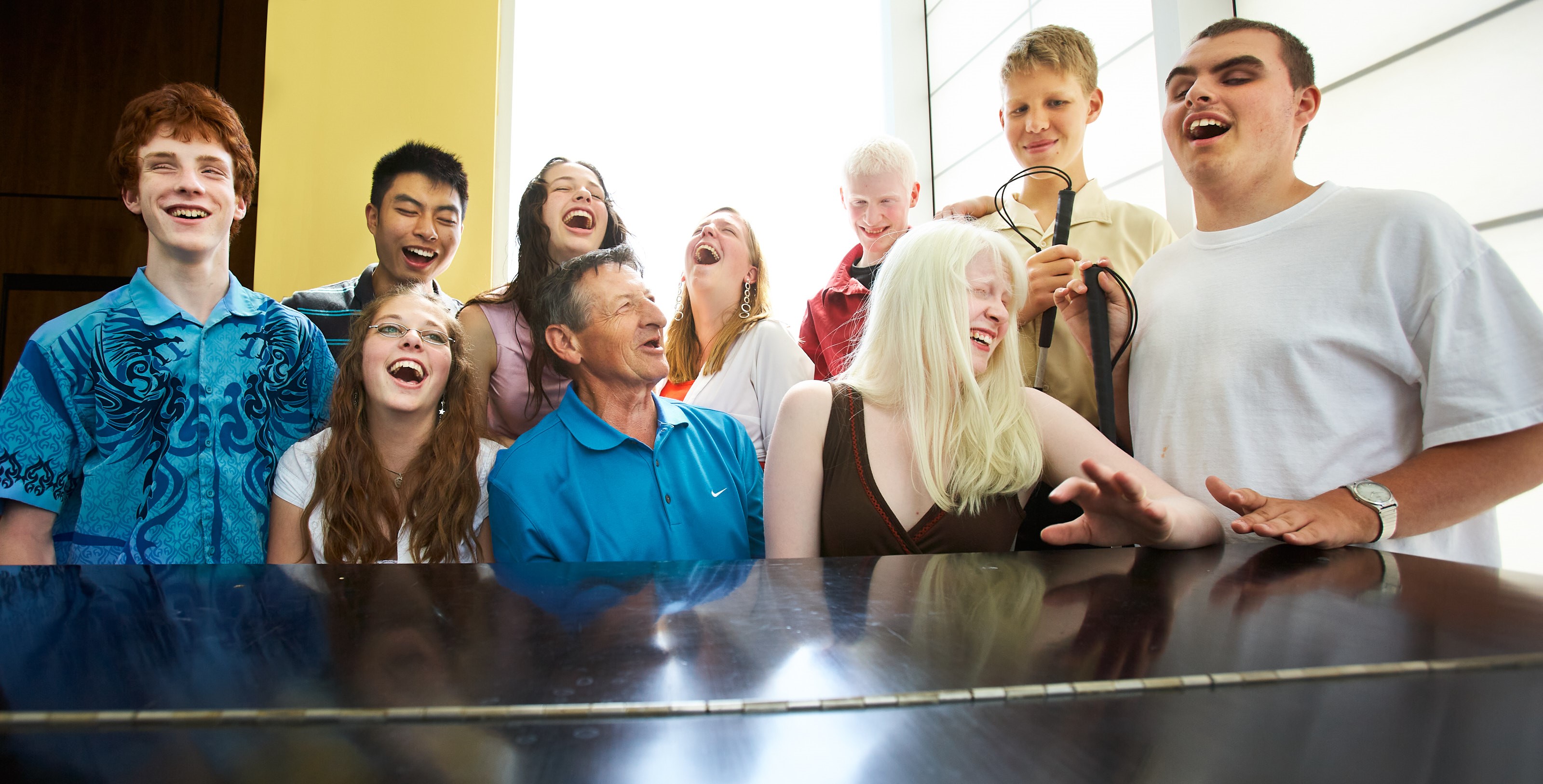 Walter Gretzky sits at a grand piano surrounded by 9 youth with sight loss. They are smiling and excitedly singing a tune while Walter plays the piano.