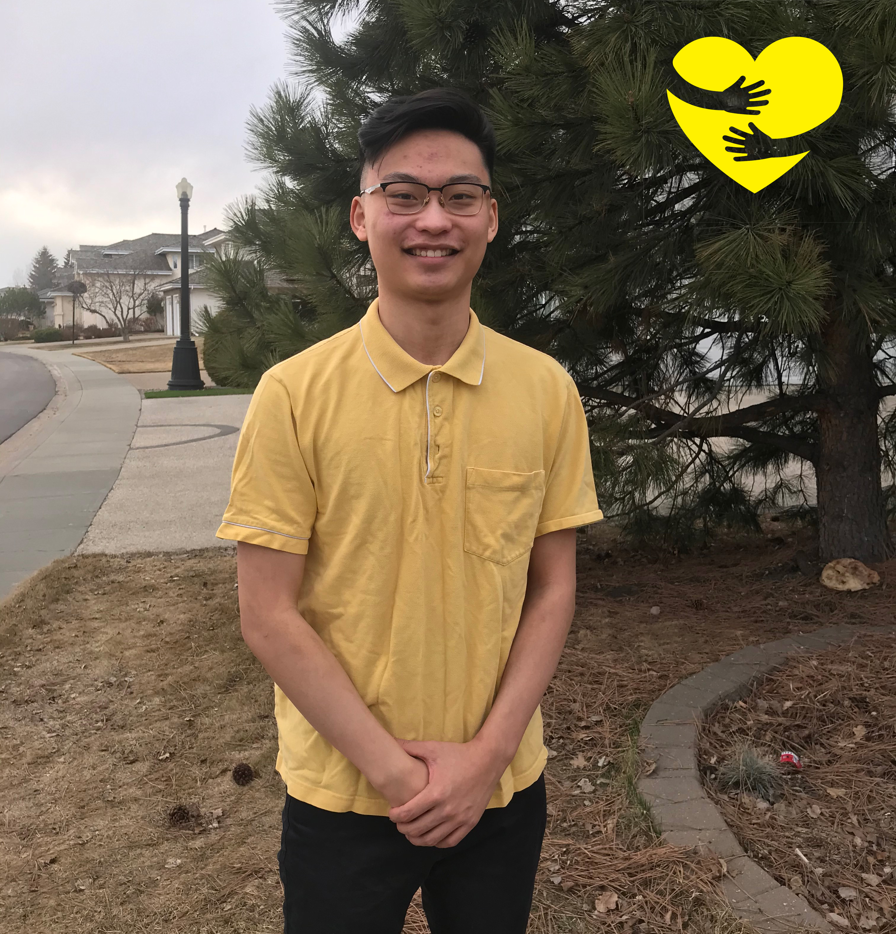 Warren, standing outside wearing a yellow shirt, smiling for the camera. A graphic of arms hugging a yellow cartoon heart can be seen in the top-right corner of the photo.