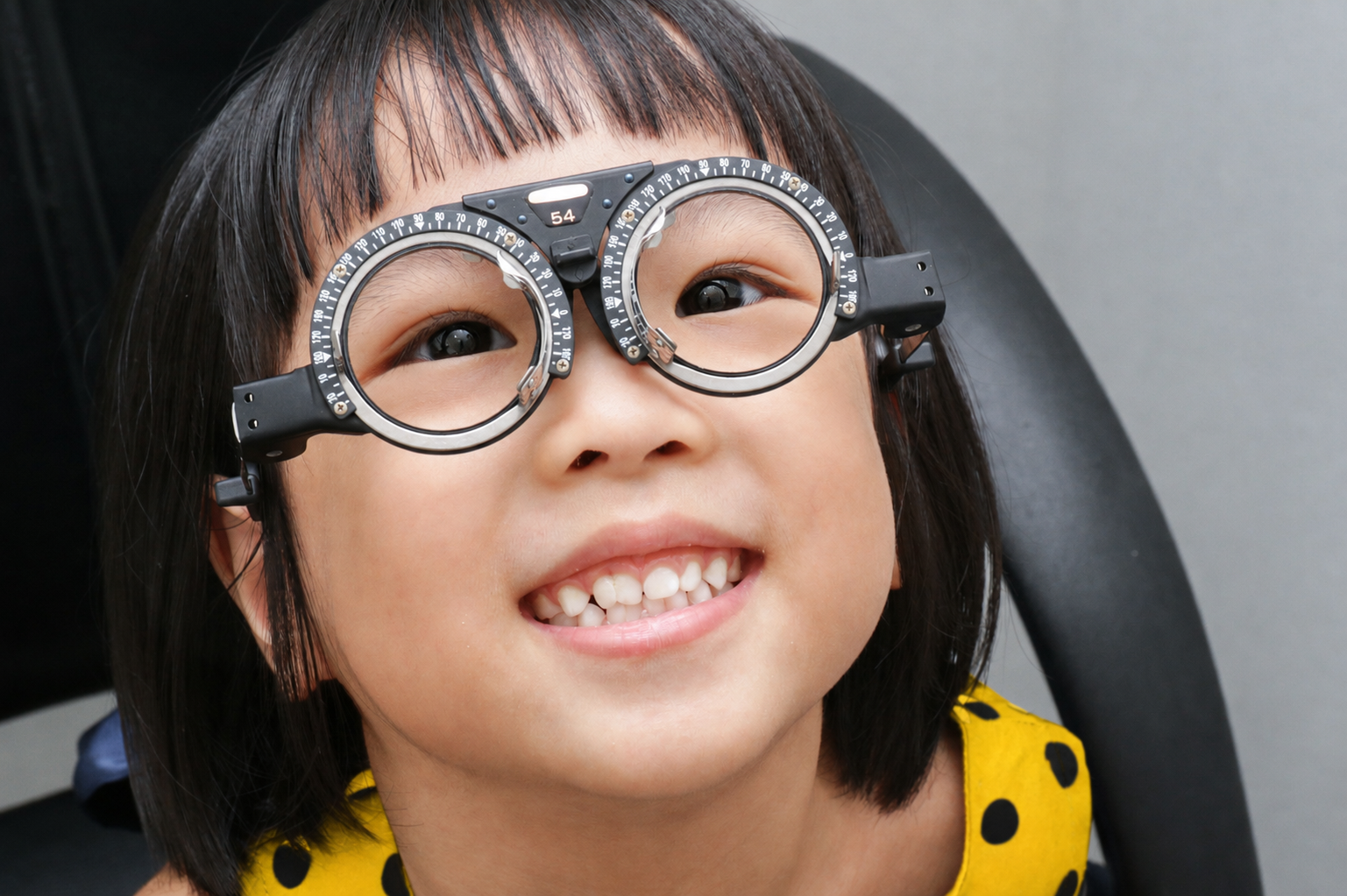 A smiling young girl with black hair and medium skin tone wears a trial frame with adjustable lenses during an eye exam.