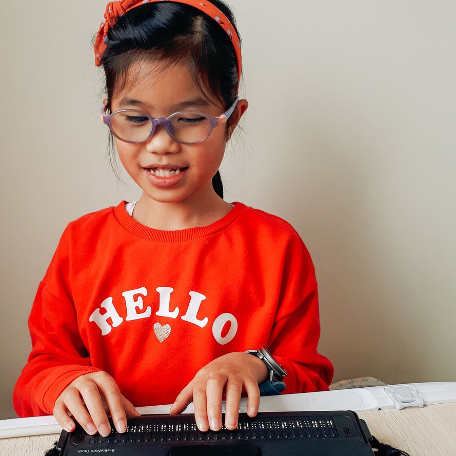A girl with glasses and medium skin uses a brailler 